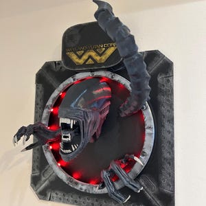 May include: A black and gray wall sculpture of an alien creature emerging from a circular opening. The creature has sharp teeth and claws. The sculpture is lit with red lights and features the Weyland-Yutani Corp logo.