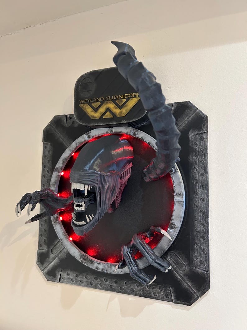 May include: A black and gray wall sculpture of an alien creature emerging from a circular opening. The creature has sharp teeth and claws. The sculpture is lit with red lights and features the Weyland-Yutani Corp logo.