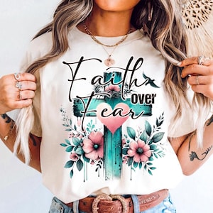May include: Cream-colored t-shirt featuring a teal cross with the words "Faith over Fear" in black script. The cross is adorned with pink flowers, green leaves, and a pink heart. A bird silhouette is also present.