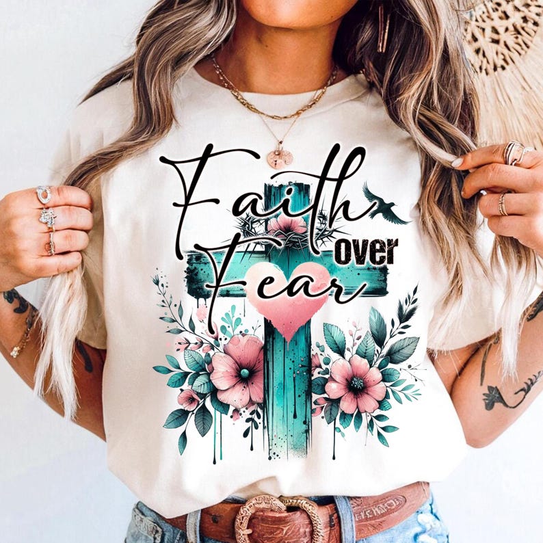 May include: Cream-colored t-shirt featuring a teal cross with the words "Faith over Fear" in black script. The cross is adorned with pink flowers, green leaves, and a pink heart. A bird silhouette is also present.
