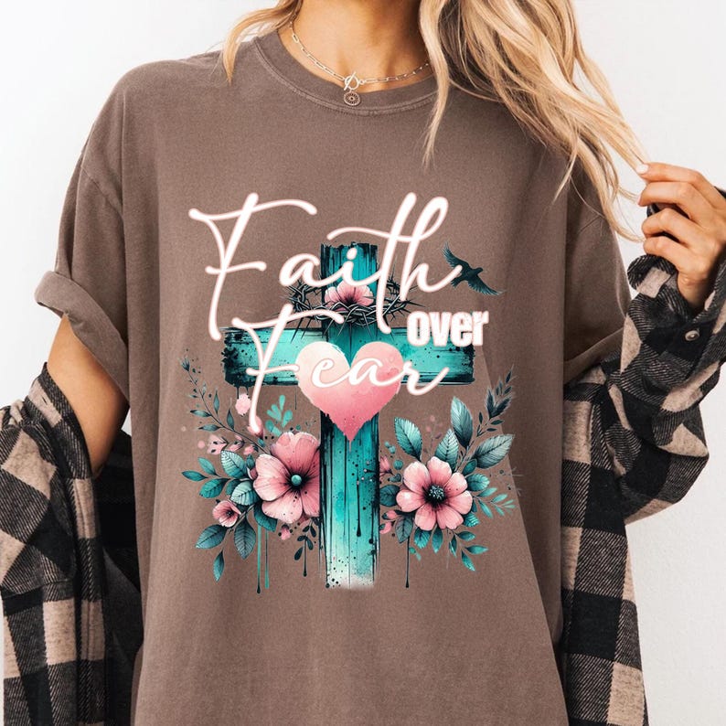 May include: Brown t-shirt with a graphic design featuring a teal cross, a pink heart, and the words "Faith over Fear." The design includes pink flowers and green leaves. A bird silhouette is also present.