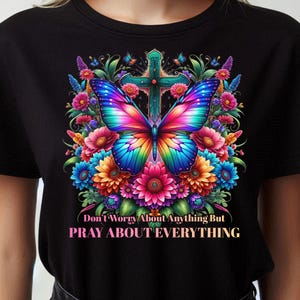 May include: Black t-shirt featuring a vibrant butterfly with rainbow wings, a teal cross, and colorful flowers. The text reads "Don't Worry About Anything But PRAY ABOUT EVERYTHING."
