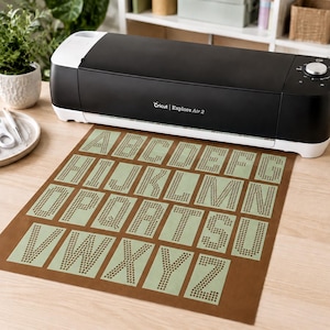 May include: A black and white Cricut Explore Air 2 electronic cutting machine sits behind a brown adhesive mat featuring a light green sheet with the letters A through Z cut out in a dotted pattern, each letter arranged in a grid on the mat.