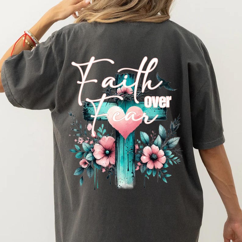 May include: Dark gray t-shirt featuring a teal cross with a pink heart and the words "Faith over Fear." The cross is surrounded by pink flowers and green leaves, creating a floral design.