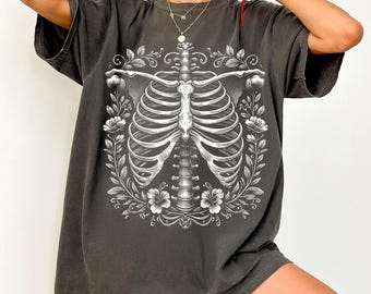 May include: Dark gray t-shirt featuring a detailed white illustration of a human rib cage surrounded by floral elements. The design is centered on the shirt, creating a striking contrast against the dark fabric. The shirt has short sleeves and a relaxed fit.