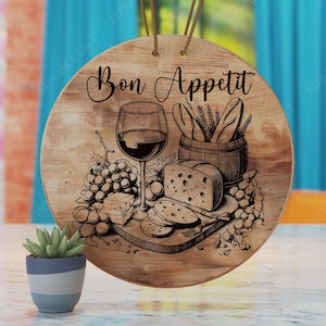 Peut inclure: Une pancarte ronde en bois avec le texte "Bon Appétit" en lettres noires. La pancarte présente une illustration dessinée à la main d'un verre de vin rouge, de raisins, de fromage, de pain et d'une fourchette sur une planche à découper en bois.