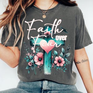 May include: Dark gray t-shirt featuring a teal cross with the words "Faith over Fear" in white script. The cross is adorned with a pink heart, flowers, and leaves. A small bird is also visible.