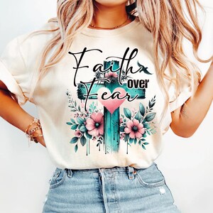 May include: Cream-colored t-shirt featuring a teal cross design with the words "Faith over Fear" in black script. The cross is adorned with pink flowers, green leaves, and a pink heart.