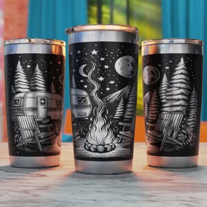May include: Three stainless steel tumblers with a black and white design of a camper, trees, a campfire, and stars. The tumblers are on a wooden surface.