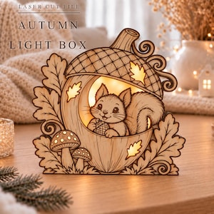 Puede incluir: Una caja de luz de madera cortada con láser que presenta un diseño en forma de bellota con una ardilla dentro, rodeada de hojas de roble y setas. El texto "LASER CUT FILE" y "AUTUMN LIGHT BOX" es visible en la parte superior.