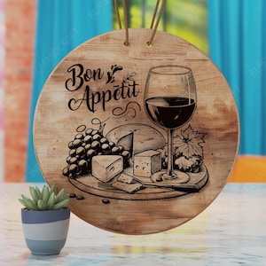 Könnte beinhalten: Ein rundes Holzschild mit dem Text "Bon Appetit" in einer dekorativen Schriftart. Das Schild zeigt eine Schwarzweißillustration eines Glases Rotwein, Trauben und Käse auf einem Holzbrett.