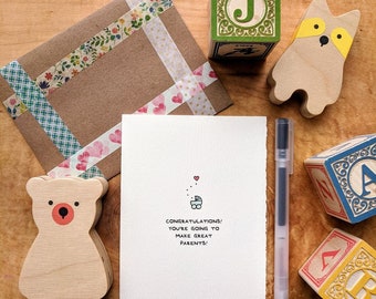 May include: A white card with the text "Congratulations! You're going to make great parents!" and a small illustration of a baby carriage with hearts above it. The card is surrounded by wooden toys, including a bear, a fox, and blocks with letters.
