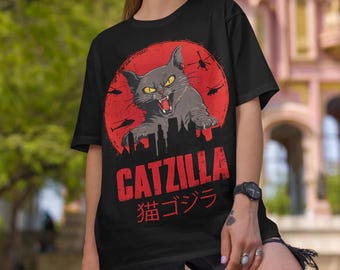 Może przedstawiać: Czarny T-shirt z czerwono-białym nadrukiem przedstawiającym kota z błyszczącymi oczami i tekstem "CATZILLA" oraz "猫ゴジラ" w języku japońskim.
