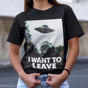 May include: Black t-shirt featuring a graphic of a UFO over a forest and the text "I WANT TO LEAVE".