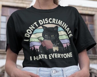 May include: Black t-shirt with a retro sunset graphic featuring a black cat reading a book and holding a cup of coffee. The text "I don't discriminate I hate everyone" is printed on the shirt.
