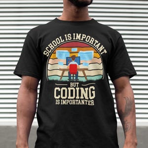 Könnte beinhalten: Schwarzes T-Shirt mit einer Retro-Grafik, auf der "Schule ist wichtig, aber Programmieren ist wichtiger" steht. Die Grafik zeigt eine Person, die an einem Computer mit mehreren Monitoren sitzt.