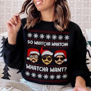 Puede incluir: Suéter navideño negro con un patrón de copo de nieve rojo, blanco y verde. El suéter presenta un gráfico de tres hombres con gorros de Papá Noel y el texto "So Whatcha Whatcha Whatcha Want?"