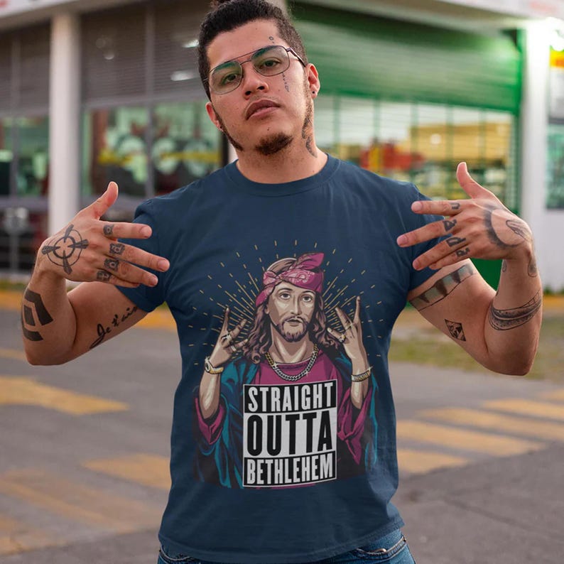 May include: A navy blue t-shirt with a graphic of Jesus wearing a bandana and a gold chain. The graphic has the text "Straight Outta Bethlehem" in a white box.