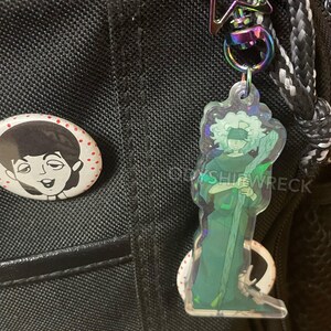 May include: A clear acrylic keychain featuring a green-toned illustration of a figure with a staff and blindfold. The keychain hangs from a rainbow-colored clasp and a star-shaped charm. A pin with a black and white illustration is also visible.