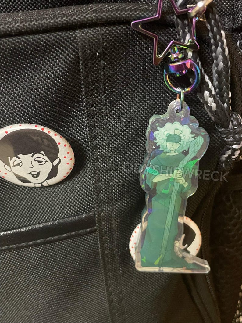 May include: A clear acrylic keychain featuring a green-toned illustration of a figure with a staff and blindfold. The keychain hangs from a rainbow-colored clasp and a star-shaped charm. A pin with a black and white illustration is also visible.