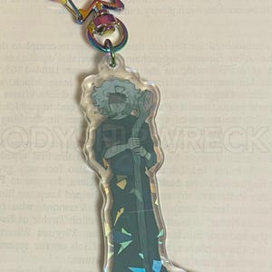 May include: A clear acrylic keychain featuring a character illustration with a blindfold and staff. The keychain has a rainbow star-shaped clasp and a circular ring. The character is dressed in dark clothing. The keychain is approximately 4 inches long.