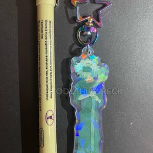 May include: A clear acrylic keychain featuring a character design with iridescent accents, attached to a star-shaped clasp. A yellow pen with black ink is next to the keychain. The pen has text that reads "Micro pigment ink for waterproof and fade proof fine lines."