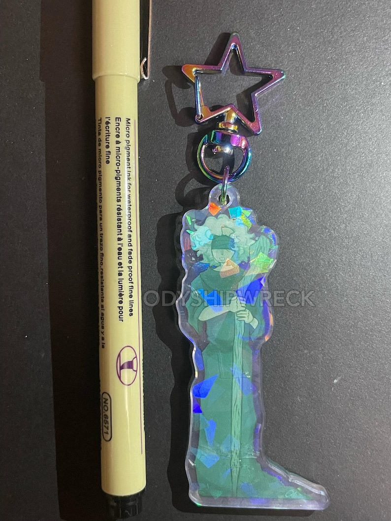 May include: A clear acrylic keychain featuring a character design with iridescent accents, attached to a star-shaped clasp. A yellow pen with black ink is next to the keychain. The pen has text that reads "Micro pigment ink for waterproof and fade proof fine lines."