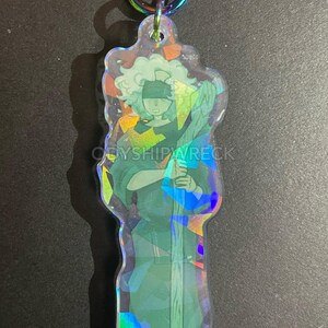 May include: A clear, iridescent keychain featuring a stylized figure in green and blue tones, holding a staff. The figure has a blindfold and curly hair. The keychain has a rainbow-colored clasp and the text "ODYSIPWRECK".