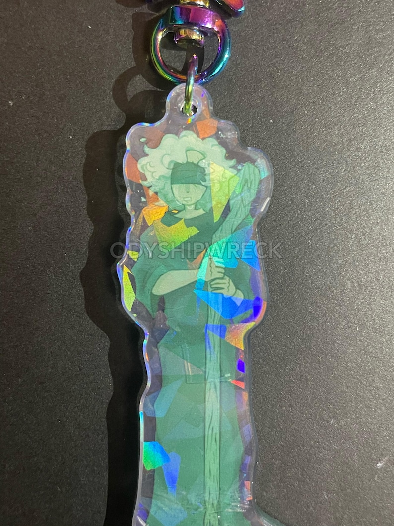 May include: A clear, iridescent keychain featuring a stylized figure in green and blue tones, holding a staff. The figure has a blindfold and curly hair. The keychain has a rainbow-colored clasp and the text "ODYSIPWRECK".