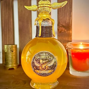 Puede incluir: Una botella redonda de vidrio amarillo dorado con un tapón dorado decorativo con alas. La botella tiene una etiqueta con texto y una ilustración detallada. Una vela encendida y un pequeño recipiente dorado están en el fondo.