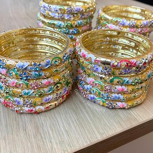 Könnte beinhalten: Mehrere Stapel goldfarbener Armreifen mit floralen und Schmuckverzierungen. Die Armreifen zeigen bunte Emailleblumen in Rosa, Blau und Weiß, akzentuiert mit kleinen, funkelnden Edelsteinen. Das Design ist aufwendig und dekorativ.