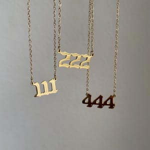以下が含まれることがあります： 111、222、444の数字ペンダントが付いた3つのゴールドカラーのネックレス。各ネックレスには繊細なチェーンが付いています。ペンダントは大胆で様式化されたフォントです。背景は無地のニュートラルカラーです。