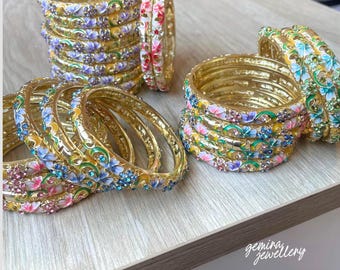 Könnte beinhalten: Eine Sammlung von Armreifen aus goldfarbenem Metall mit farbenfrohen Emaille-Blumenmustern und eingebetteten Strasssteinen. Die Armbänder sind in drei Stapeln angeordnet und zeigen detaillierte, bemalte Blütenblätter in Rosa, Blau und Grün.