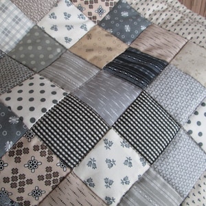 May include: Close-up of a patchwork quilt with a variety of patterned squares. The squares feature designs in shades of gray, white, and tan, including polka dots, stripes, and floral prints. The quilt has a diamond pattern.