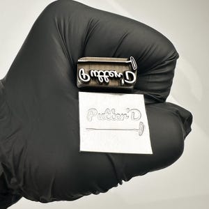 May include: A metal stamp with the text "Putter'D" in a cursive font. The stamp is shown with an impression of the text on a square piece of metal. The stamp is held by a gloved hand.