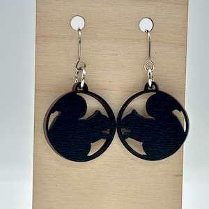 May include: A pair of black squirrel-shaped earrings. Each earring features a circular design with a squirrel silhouette cut out of the center. The earrings hang from silver-colored fish hook ear wires. The earrings are displayed on a light-colored wooden surface.