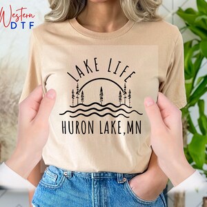 May include: A tan t-shirt with black text that reads "Lake Life Huron Lake, MN". The text is stylized with a sun and trees.