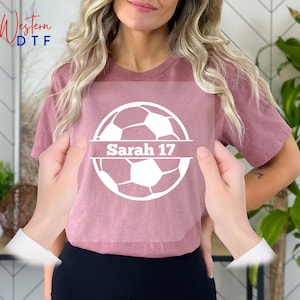 May include: A pink t-shirt with a white soccer ball graphic and the text "Sarah 17" on a white banner.