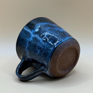May include: A handmade ceramic mug with a dark blue, glossy glaze. The mug features a rounded body, a sturdy handle, and a textured, brown base. The blue glaze has lighter, swirling patterns, creating a unique, artistic appearance. This mug is perfect for coffee or tea.