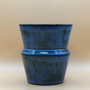 May include: A handmade ceramic vase in shades of blue and green. The vase has a unique shape with a flared top and a narrower midsection. The glaze has a speckled appearance, adding to its rustic charm. This decorative piece is perfect for displaying flowers or as a standalone art object.