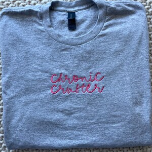May include: A heather gray t-shirt with the text "Chronic Crafter" in pink script. The shirt is folded neatly, showcasing the design prominently. The fabric appears soft and comfortable.