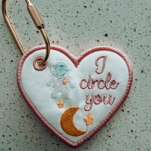May include: Heart-shaped embroidered keychain with a gold-colored clasp. The white fabric features an astronaut, crescent moon, stars, and the text "I circle you" in pink thread. The heart is outlined in pink.