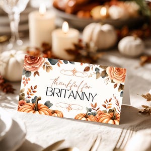 Könnte beinhalten: Eine Tischkarte zum Thema Thanksgiving mit dem Text "thankful for BRITANNY". Die Karte zeigt Aquarellillustrationen von Kürbissen, Rosen und Herbstlaub in Orange, Braun und Grün. Sie liegt auf einer weißen Tischdecke.