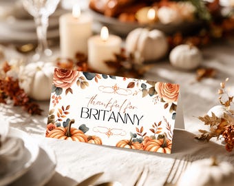 Könnte beinhalten: Eine Tischkarte zum Thema Thanksgiving mit dem Text "thankful for BRITANNY". Die Karte zeigt Aquarellillustrationen von Kürbissen, Rosen und Herbstlaub in Orange, Braun und Grün. Sie liegt auf einer weißen Tischdecke.