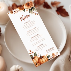 K&ouml;nnte beinhalten: Eine Thanksgiving-Men&uuml;karte mit floralem und K&uuml;rbis-Design. Das Men&uuml; bietet eine Vorspeise, einen Hauptgang, Beilagen und ein Dessert. Die Karte liegt auf einem wei&szlig;en Teller mit Kerzen und K&uuml;rbissen im Hintergrund.