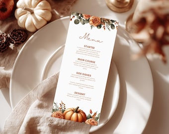 Könnte beinhalten: Eine Menükarte mit floralen und Kürbis-Illustrationen. Die Karte zeigt das Wort "Menu" in eleganter Schrift. Das Menü listet eine Vorspeise, einen Hauptgang, Beilagen und ein Dessert auf. Die Karte liegt auf einem weißen Teller mit einer Leinen-Serviette.