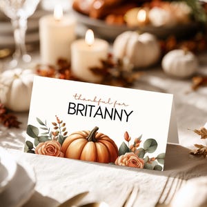 Könnte beinhalten: Eine Tischkarte für Thanksgiving mit dem Text "thankful for BRITTANY". Die Karte zeigt ein Aquarelldesign mit einem Kürbis, Rosen und Grünzeug. Die Karte liegt auf einem Tisch mit Kerzen und Kürbissen.