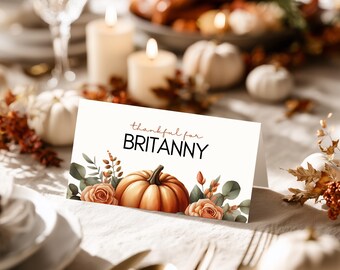 Puede incluir: Una tarjeta de mesa de Acción de Gracias con el texto "thankful for BRITTANY". La tarjeta presenta un diseño de acuarela de una calabaza, rosas y vegetación. La tarjeta está colocada sobre una mesa con velas y calabazas.