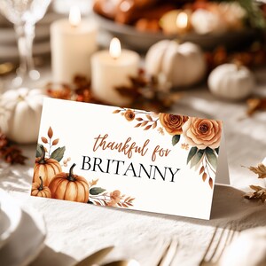 May include: A folded place card with the text "thankful for BRITTANY" in brown script. The card is decorated with watercolor pumpkins, roses, and fall foliage. The card is set on a white tablecloth with candles and pumpkins in the background.