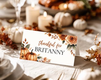 Könnte beinhalten: Eine gefaltete Tischkarte mit dem Text "thankful for BRITTANY" in brauner Schrift. Die Karte ist mit Aquarellkürbissen, Rosen und Herbstlaub verziert. Die Karte liegt auf einer weißen Tischdecke mit Kerzen und Kürbissen im Hintergrund.